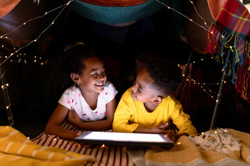 Happy african american siblings lying in makeshift tent, using tablet © wavebreak3