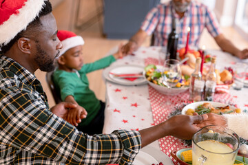 Happy multi generation family wearing santa hats, praying together, having christmas meal