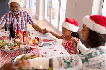 Happy multi generation family wearing santa hats, praying together, having christmas meal