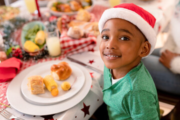 Happy african american boy wearing santa hat, sitting at table, looking at camera