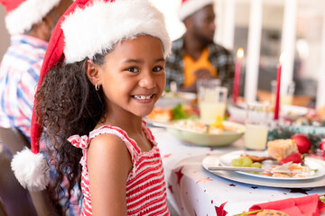 Happy african american girl wearing santa hat sitting at christmas table