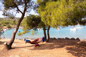 Person lying in colorful hammock in the shade of pine trees on the Kosirina beach, Murter island, Croatia, and boats moored in a bay