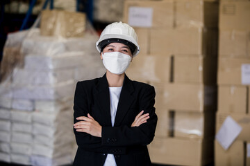 portrait of manager women in a black suit wearing a disposable face mask and helmet standing cross arms looking confident in a factory warehouse, logistic industry concept.