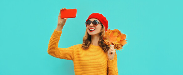 Autumn portrait of happy smiling young woman taking selfie by smartphone with yellow maple leaves wearing a sweater, french beret on blue background