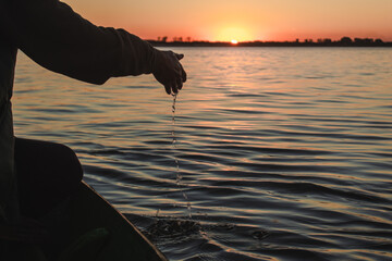 Hand of a young man in a boat touching the sea with sunset in the background.
