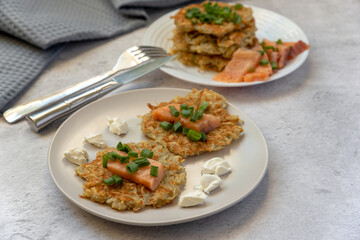 Potato pancakes with slices of lightly salted salmon with cottage cheese and green onions on plates with cutlery and a gray cloth on a gray background