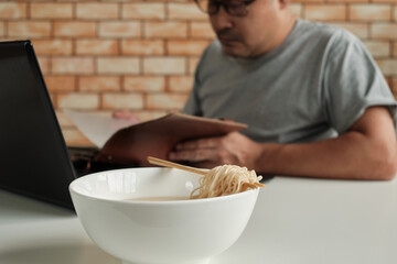 Thai male worker busy working with laptop, use chopsticks to hastily eat instant noodles during office lunch's break, because quick, tasty, and cheap. Over time Asian fast food, unhealthy lifestyle.