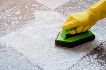 Fototapeta na wymiar Human hands wearing yellow rubber gloves are using a green color plastic floor scrubber to scrub the tile floor with a floor cleaner.