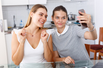 Two cheerful young girls taking selfie in kitchen interior