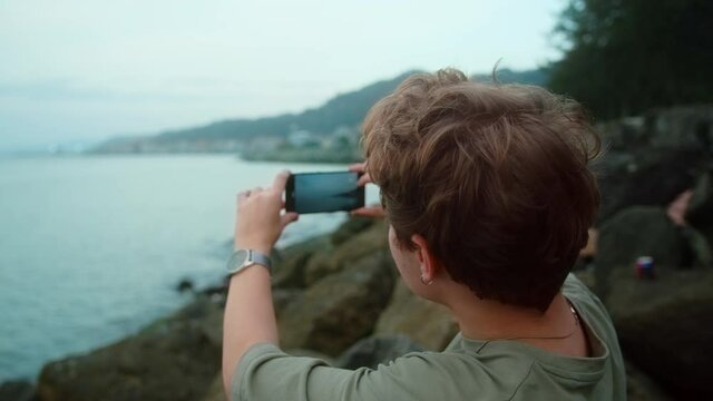 Young  blonde boy watching sunset and sea over bay from top of the stone, he takes panorama photos or record video on phone