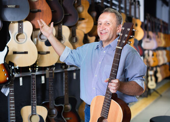 Adult glad smiling guitarist is standing with acoustic guitar in music store.