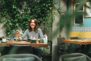 Young smiling brunette woman in casual clothes using social media on mobile phone sitting at the green cafe