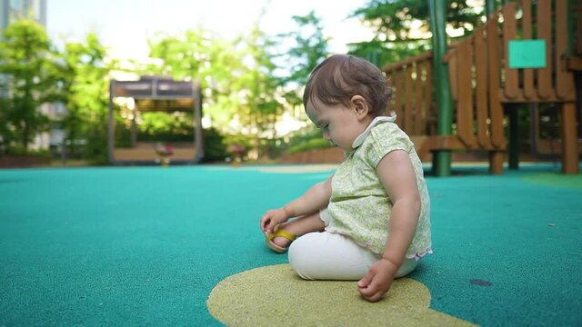Lonely One-year-old cute little baby girl sitting on the floor of outdoor kid's playground alone and pick up small stone from the ground