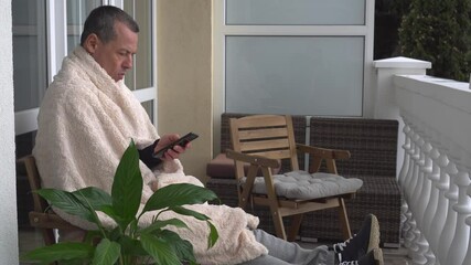 A middle-aged man relaxes in autumn at home on the balcony, reads a book, wrapped comfortably in a warm winter blanket. Self-isolation lockdown quarantine lifestyle during Coronavirus pandemic