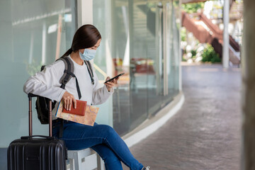 Travel. A woman in a white long-sleeved shirt and jeans, wearing a mask. sit on the chair and read a map in her hand while suitcase and backpack are placed on a side