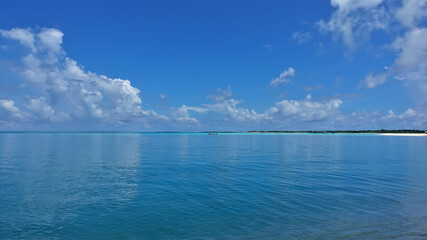 The aquamarine ocean is calm. There are picturesque cumulus clouds in the azure sky. A tropical island is visible in the distance. Reflection on the water surface. Maldives