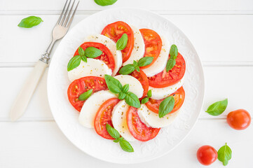 Classic caprese salad with mozzarella cheese, tomatoes and basil on a white plate on a wooden background. Top view, copy space.