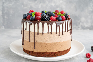 Three chocolate mousse cake with chocolate glaze, blueberries, raspberries and blackberries on top on a white plate on a gray concrete background. Cake with three layers of different taste. Copy space