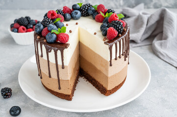 Three chocolate mousse cake with chocolate glaze, blueberries, raspberries and blackberries on top on a white plate on a gray concrete background. Cake with three layers of different taste. Copy space