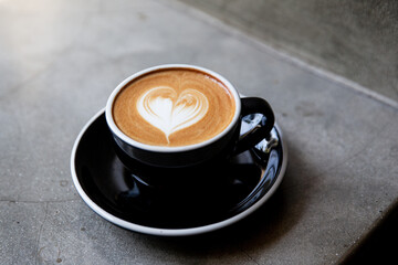 Black cup of cappuccino with latte art of heart shape on saucer on concrete background.