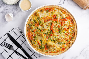 Baked french vegetable pie quiche filled with carrots, peas and cheese on a marble surface, cracked egg shell, flour, cream, top view