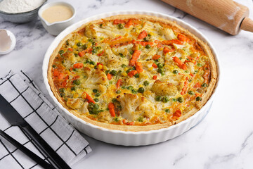 Baked french vegetable pie quiche filled with carrots, peas and cheese on a marble surface, cracked egg shell, flour, cream