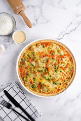 Baked french vegetable pie quiche filled with carrots, peas and cheese on a marble surface, cracked egg shell, flour, cream, top view