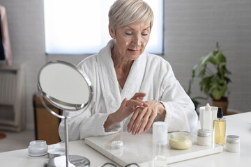 An older woman with short blonde hair in a white bathrobe sits at the table and puts cream on her hands inside the living room
