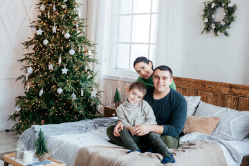 Happy beautiful family with a small son sitting on the couch in the house on the background of the Christmas tree - 458667067
