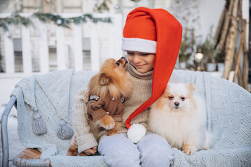 A little boy dressed in a red Santa hat holds two Spitz puppies in his arms. The child sits with dogs on the background of a white winter house. Christmas present for kid - 458667472