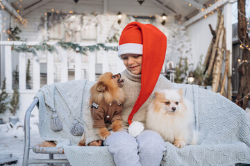 A little boy dressed in a red Santa hat holds two Spitz puppies in his arms. The child sits with dogs on the background of a white winter house. Christmas present for kid - 458667495