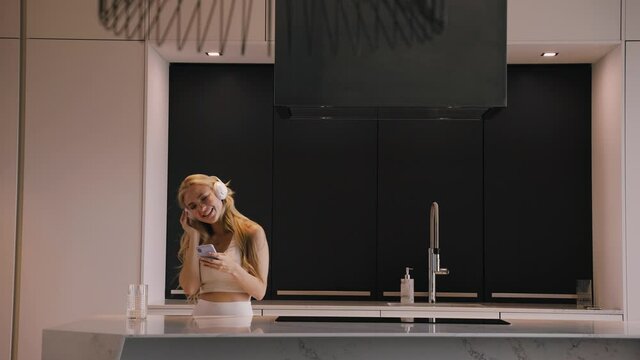 A young blonde woman with a phone in her hands and headphones in the kitchen happily listens to music, dancing, chat, reads the news.