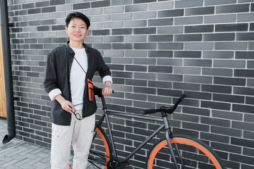 Fototapeta na wymiar Horizontal medium long shot of young Asian woman with short hair wearing fashionable casual outfit standing with bicycle against gray brick building smiling at camera
