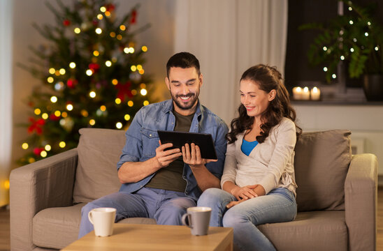 holidays, technology and people concept - happy couple using tablet computer at home in evening over christmas tree lights on background