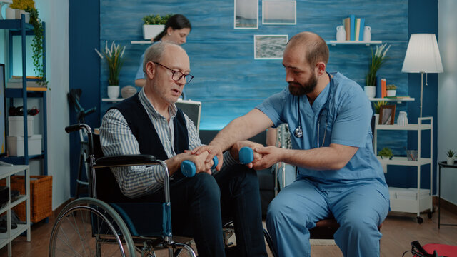 Nurse helping disabled man to exercise with dumbbells for physical activity and recovery in nursing home. Medical assistant giving assistance to old patient with chronic disease in wheelchair.