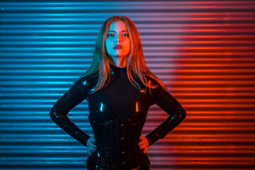 Red-haired girl in latex, a black top and tight pants, on a metallic background illuminated with red and blue LEDs, posing with her hands on her hips