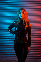 Red-haired girl in latex, a black top and tight pants, on a metallic background illuminated with red and blue LEDs - obrazy, fototapety, plakaty