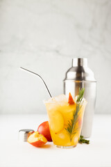Cooking of fresh cold peach drink in glass with ice cubes, green rosemary, fruit slices, ingredients, silver shaker in soft light white kitchen interior, vertical.