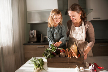 mom and child daughter  in a linen apron with a box  of fresh garden vegetables in the kitchen....