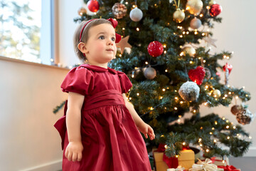 winter holidays and childhood concept - lovely baby girl in red dress over christmas tree at home