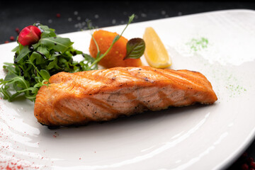 Fried salmon steak with herbs and lemon on a white plate on a dark background
