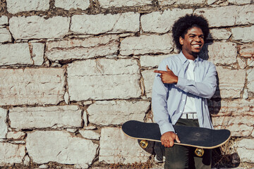 Fototapeta na wymiar Smiling young African American student leaning against a wall with a skate and pointing the copy space