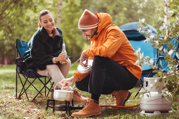 camping, tourism and travel concept - happy couple drinking beer and cooking food in pot on tourist gas burner at tent camp © Syda Productions