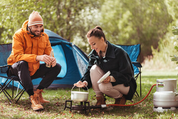 camping, tourism and travel concept - happy couple drinking beer and cooking food in pot on tourist...