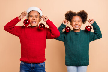 Photo of two funny cute kids brother sister hold tree baubles wear x-mas hat jumper isolated beige color background