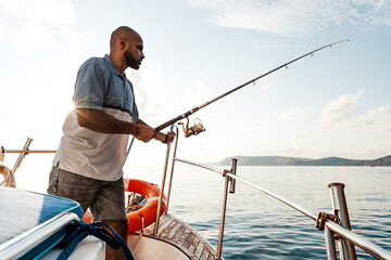 Young african american man standing with fishing rod on a sailboat fishing in open sea on sunset - Powered by Adobe