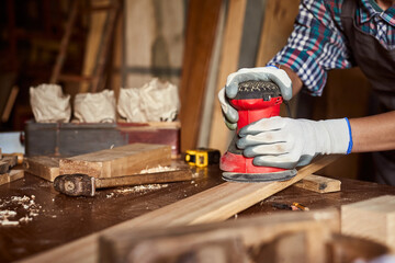 Female carpenter grinding wood with sandpaper in carpentry or diy workshop. Electric sander working in carpentry. Girl polishes wooden board with electric orbital sander.