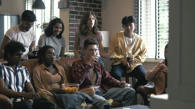 Happy excited diverse friends students watching movie, commenting a film. Multiethnic positive young people watching TV, sitting hanging out together eating snacks in the living room