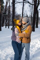 Fototapeta na wymiar Smiling couple in warm clothes looking at each other while hugging in winter park