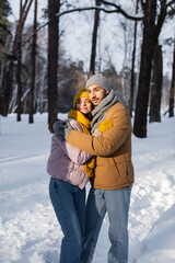 Fototapeta na wymiar Young couple in winter outfit embracing while standing in sunlight and snow